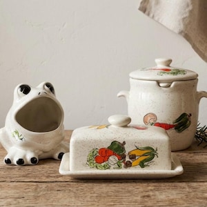 May include: A ceramic frog-shaped container, a rectangular butter dish with a lid, and a lidded jar, all featuring a speckled off-white finish. The butter dish and jar are decorated with colorful hand-painted illustrations of various vegetables.