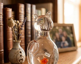 May include: A clear glass cat figurine with an orange fish inside. The cat has a rounded body, a tail, and pointed ears. The figurine is sitting on a wooden surface, with books and a small vase in the background.