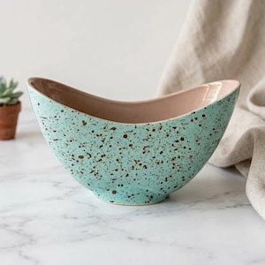 May include: A curved, boat-shaped ceramic bowl featuring a light mint green exterior with a dark brown speckled pattern. The interior of the bowl has a smooth, solid light beige finish, and the overall design has a wide, open top edge.