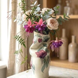 May include: A cream-colored ceramic vase with a twisted, organic shape, decorated with painted fuchsia flowers and green leaves. The vase holds a bouquet of fresh fuchsia flowers, pale pink roses, and green eucalyptus branches.