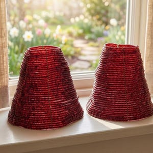 May include: Two red beaded lampshades with a conical shape sit on a windowsill. The shades have a textured surface and are made of small, closely-knit beads. The background shows a garden with flowers and a window.