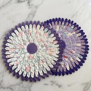 May include: Two handmade fabric coasters. One coaster features a white and pastel floral pattern with a purple center and border. The other coaster is a solid purple with a purple center. Both are circular and made of folded fabric.