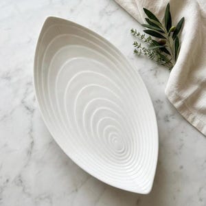 May include: White, leaf-shaped serving platter with concentric circle design. The ceramic dish is on a marble surface, with a linen cloth and sprigs of greenery in the background. A decorative piece for serving food.