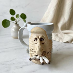 May include: A ceramic mug featuring a sculpted human face with a prominent nose, closed eyes, and a textured beard area. The mug has a light blue glazed rim and handle, with three white cylindrical objects protruding from the mouth area.