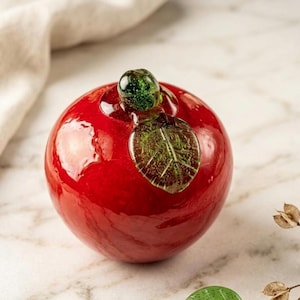 May include: A glossy red glass apple with a green stem and leaf. The decorative apple is a vibrant piece of art, perfect for home decor. The apple is a bright, eye-catching accent piece.