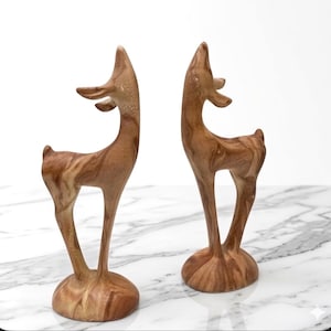 May include: Two wooden deer sculptures with slender legs and curved necks. The deer are carved from wood with a natural grain pattern, standing on round bases. The sculptures are a warm brown color.