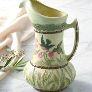 May include: A light green ceramic pitcher with a curved spout and a handle. The pitcher is decorated with images of hummingbirds and pink flowers. The base and top of the pitcher have a textured design. The handle has a brown accent.