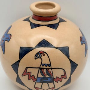 May include: A round, beige ceramic vase with a small opening and a red band near the top. The vase is decorated with a stylized eagle design in blue and red, surrounded by dark blue starburst shapes.