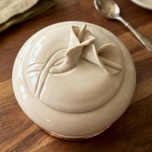 May include: A cream-colored ceramic sugar bowl with a lid featuring a raised floral design. The bowl has a copper-colored base and sits on a wooden surface. A spoon and a small plate are visible in the background.