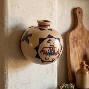 May include: A beige, round ceramic vase with a small opening and a flat back for wall mounting. The vase features a Native American-inspired design with a central eagle motif in red, blue, and black, surrounded by a starburst pattern. A star and other geometric shapes are also painted on the vase.