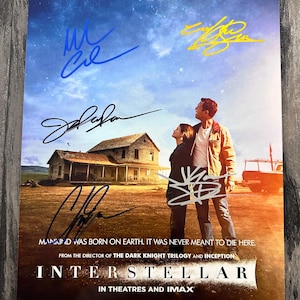 May include: A movie poster for "Interstellar" featuring a landscape with a house and two people looking up at the sky. The poster has multiple signatures and the text "Mankind was born on Earth. It was never meant to die here."