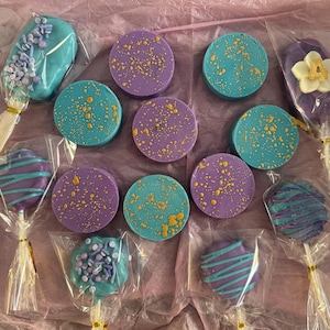 May include: An assortment of decorated treats, including teal and purple cookies, cake pops, and pretzel rods. The cookies have gold speckles, and the cake pops and pretzel rods are decorated with sprinkles and icing. All treats are individually wrapped.