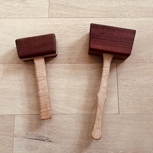 May include: Two wooden mallets with contrasting colours. The mallet heads are a dark reddish-brown, while the handles are a light, natural wood. The mallets are lying on a light-coloured wooden surface.