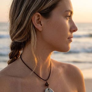 Puede incluir: Un collar con un colgante claro y texturizado suspendido de un cordón marrón oscuro. Una cuenta oscura se encuentra encima del colgante. El fondo muestra una escena de playa con el mar y una puesta de sol.