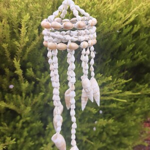 May include: A hanging mobile crafted from numerous small white and cream-coloured seashells. The structure features a circular wire frame adorned with shells, from which multiple vertical strands of various shell types dangle downwards.