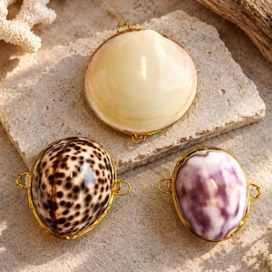 May include: Three decorative shell charms with gold-toned metal rims and small loops on each side. One shell is smooth and cream-coloured, one features a brown spotted pattern, and the third has a marbled purple and white surface texture.