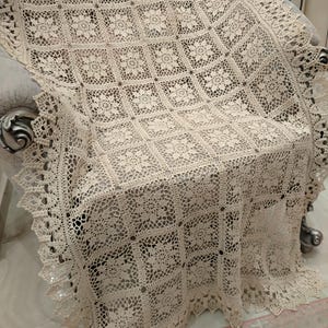 Puede incluir: Una manta de ganchillo color crema drapeada sobre una silla. La manta presenta un patrón cuadrado repetitivo con motivos florales y un borde festoneado. El intrincado diseño calado añade una textura delicada al textil.