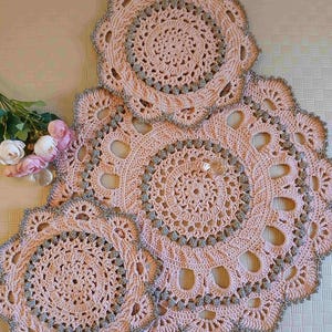 Peut inclure: Un ensemble de trois napperons ronds en crochet rose clair, présentant des motifs ajourés complexes et une bordure décorative rehaussée de fil argenté métallique. Chaque pièce affiche un design circulaire central avec des détails argentés.