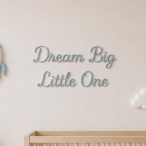 May include: A wooden crib with a light blue blanket, a blue teddy bear, a star-shaped lamp, and a small house figurine on a shelf. The wall features a blue rocket ship, three white clouds, and the script text "Dream Big Little One" in gray.