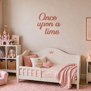 May include: A white toddler bed with a pink crown detail, featuring a pink quilted blanket, a pink throw with pom-poms, a pink pillow, and a white pillow with the text "little prince". The wall displays the pink text "Once upon a time".
