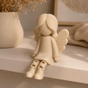 Könnte beinhalten: Eine cremefarbene Figur eines gesichtslosen Engels mit langem Haar, der ein schlichtes Kleid trägt und bewegliche Beine hat, die über die Kante hängen. Der Engel hat markante Flügel auf dem Rücken und besteht aus einem glatten, matten Material.