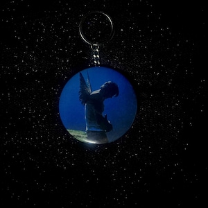 May include: A round keychain featuring a silhouette of a figure with angel wings against a blue background. The figure is bent over, with the wings spread. The keychain has a silver ring and chain.