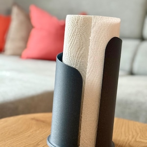 Könnte beinhalten: Ein schwarzer Papierhandtuchhalter mit einer Rolle weißer Papierhandtücher. Der Halter ist zylindrisch mit einer gebogenen Öffnung an der Vorderseite. Der Halter steht auf einem Holztisch.