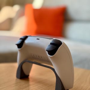 Könnte beinhalten: Ein weiß-schwarzer Videospiel-Controller auf einem dunkelgrauen Ständer. Der Controller hat schwarze Thumbsticks und Tasten. Der Ständer hält den Controller. Der Hintergrund umfasst einen Holztisch und ein verschwommenes Wohnzimmer. Für Videospiele.