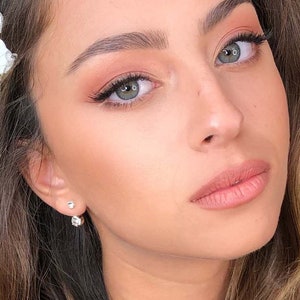 May include: A woman with long brown hair and a white flower in her hair. She is wearing natural makeup with a light pink lip color.