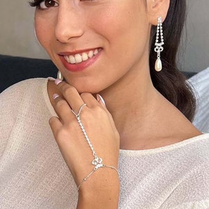 May include: A woman wearing a white top with a silver chain bracelet and dangling pearl earrings with rhinestones.