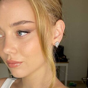 May include: A woman with blonde hair and light makeup, wearing a white tank top. Her hair is styled in a loose updo and she is wearing small diamond earrings.