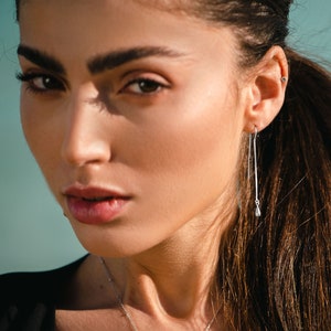 Puede incluir: Primer plano de la cara de una mujer con el pelo largo y castaño. Lleva un collar de plata y pendientes de gota de plata. Sus ojos son marrones y sus labios están ligeramente entreabiertos.