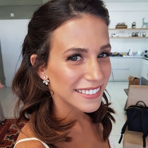 May include: A woman with long brown hair wearing a white top and gold earrings with clear stones. She is smiling and looking at the camera.