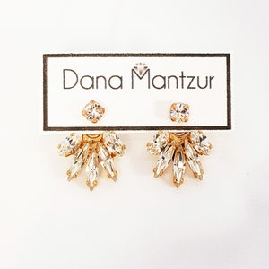 May include: Gold earrings with a cluster of clear crystals in a fan shape. The earrings are on a white card with the text "Dana Mantzur" printed on it.
