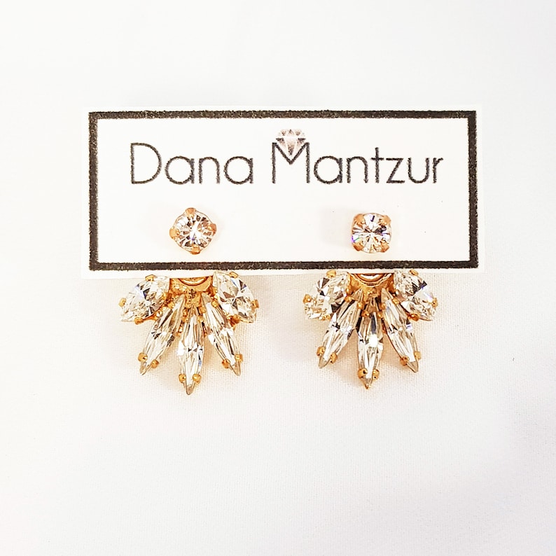 May include: Gold earrings with a cluster of clear crystals in a fan shape. The earrings are on a white card with the text "Dana Mantzur" printed on it.