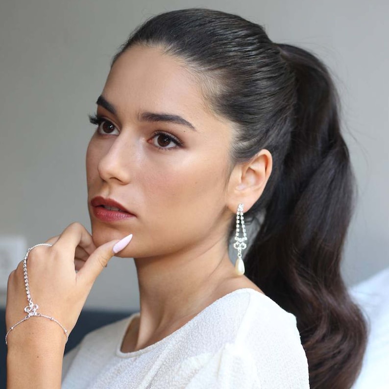 May include: A woman with long dark hair pulled back in a ponytail wears a white top and silver jewelry. She is wearing a silver bracelet with a small crystal design and dangling silver earrings with a pearl drop.