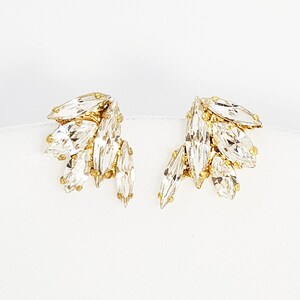 May include: A pair of gold earrings featuring clear crystal marquise-shaped stones. The stones are clustered together and set in a gold setting.