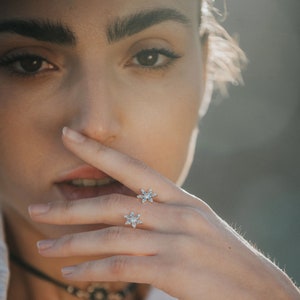 May include: Two gold rings with snowflake designs featuring clear cubic zirconia stones. The rings are worn on the index and middle fingers of a person's hand.