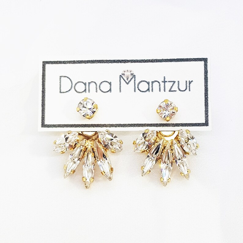 May include: Gold earrings with a cluster of clear crystals in a fan shape. The earrings are displayed on a white card with the text "Dana Mantzur" printed on it.