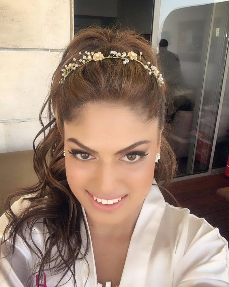 May include: A woman with long brown hair wearing a white robe and a gold headband with small white flowers and rhinestones.