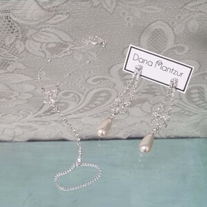 May include: A silver bracelet with a pearl drop and matching earrings with pearl drops. The earrings and bracelet are adorned with rhinestones. The jewelry is displayed on a white card with the text "Dana Mantzur".