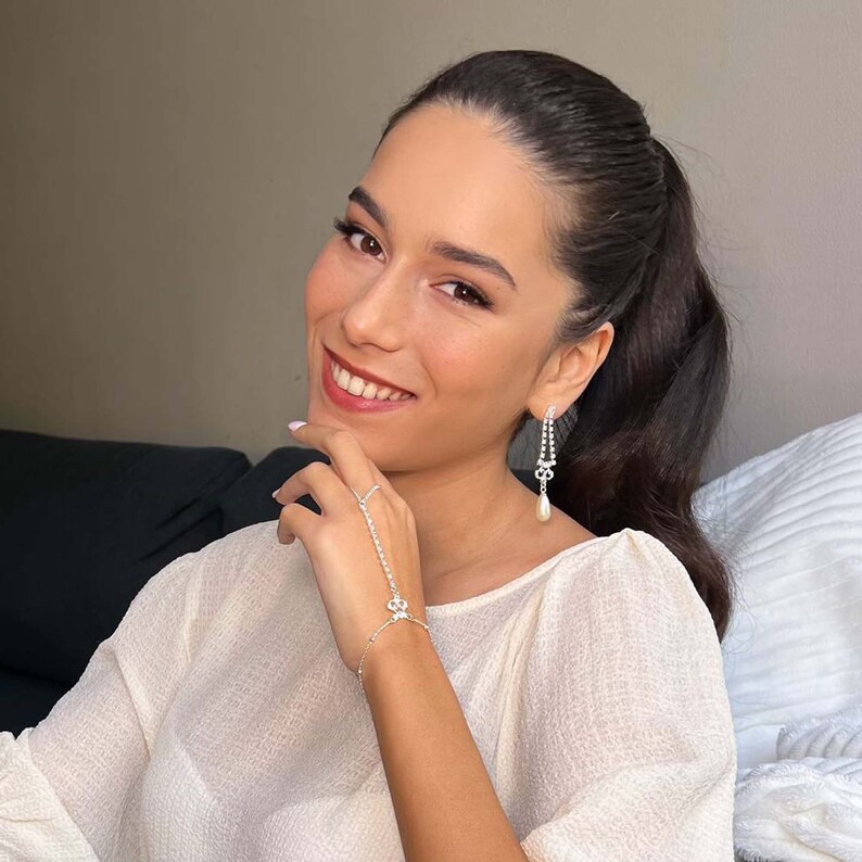 May include: A woman wearing a white top with a silver rhinestone bracelet and matching earrings with a pearl drop.