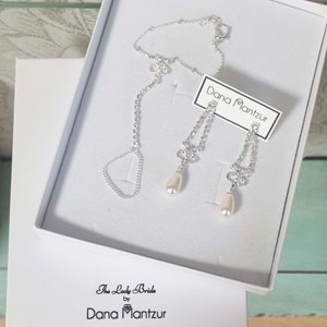 May include: A silver necklace and earrings set with a pearl drop pendant. The jewelry is in a white box with the text "The Lady Bride by Dana Mantzur" on the bottom and "Dana Mantzur" on a tag.