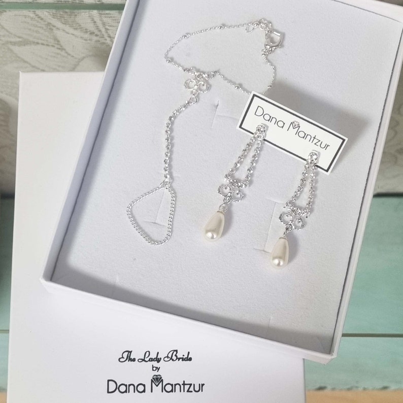 May include: A silver necklace and earrings set with a pearl drop pendant. The jewelry is in a white box with the text "The Lady Bride by Dana Mantzur" on the bottom and "Dana Mantzur" on a tag.