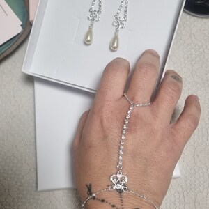 May include: A silver bracelet with a crystal design and a matching pair of silver earrings with pearl drops. The earrings are displayed in a white box with the text "Dana Mantzur" on the label.