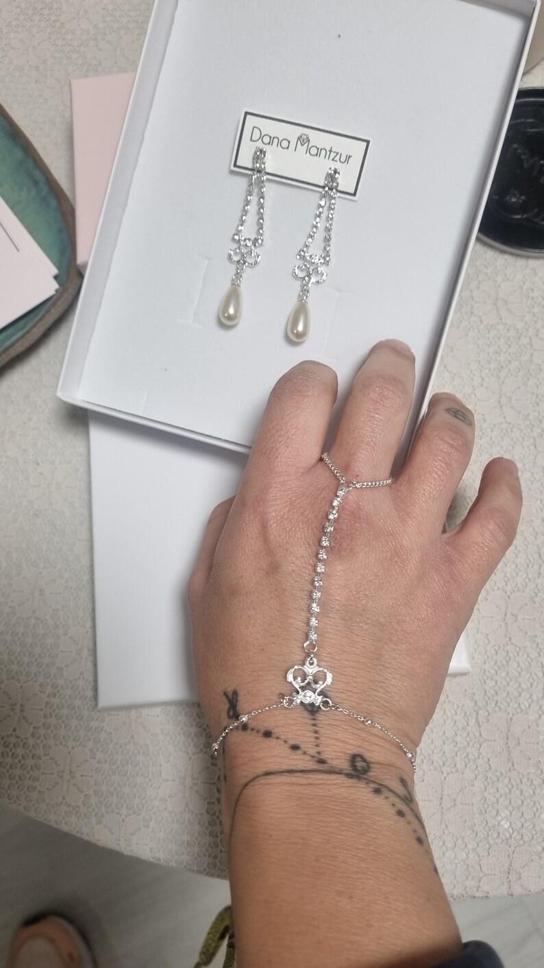 May include: A silver bracelet with a crystal design and a matching pair of silver earrings with pearl drops. The earrings are displayed in a white box with the text "Dana Mantzur" on the label.
