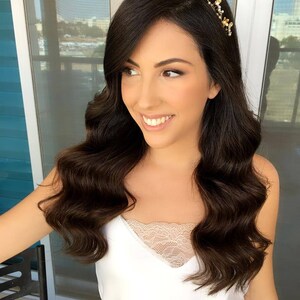 May include: A woman with long, dark brown hair styled in loose waves wears a gold headband with small white beads and gold accents. She is wearing a white tank top with a lace trim.