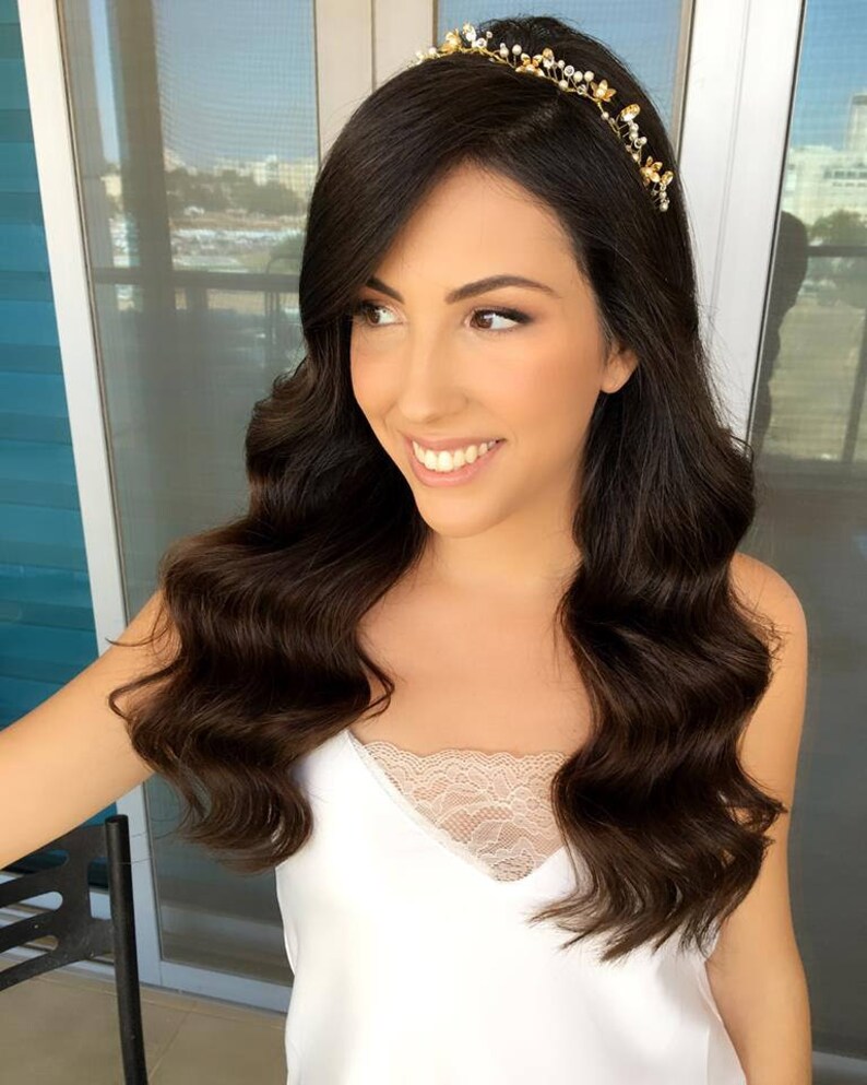 May include: A woman with long, dark brown hair styled in loose waves wears a gold headband with small white beads and gold accents. She is wearing a white tank top with a lace trim.