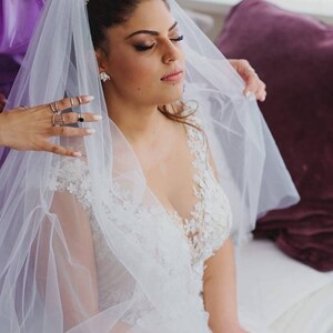 May include: A bride wearing a white lace wedding dress with a veil being placed on her head by a bridesmaid. The bride is wearing a gold headband with small flowers.