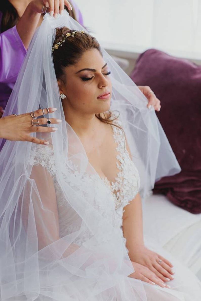 May include: A bride wearing a white lace wedding dress with a veil being placed on her head by a bridesmaid. The bride is wearing a gold headband with small flowers.